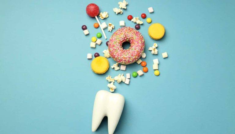A model tooth on a blue background beneath a scattering of doughnut, lollipop, macarons, marshmallows and popcorn
