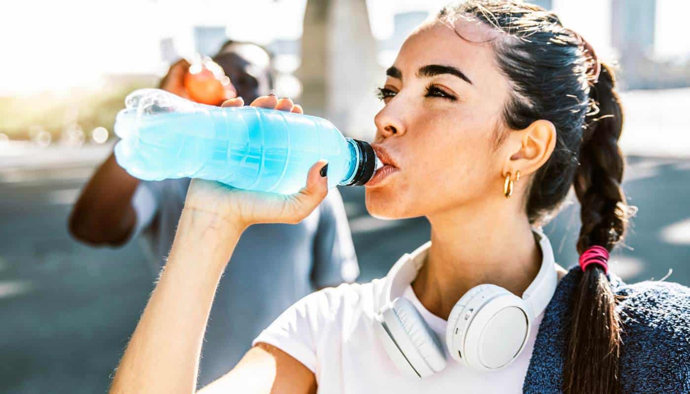 A woman in sportswear drinking a blue sports drink from a bottle outdoors