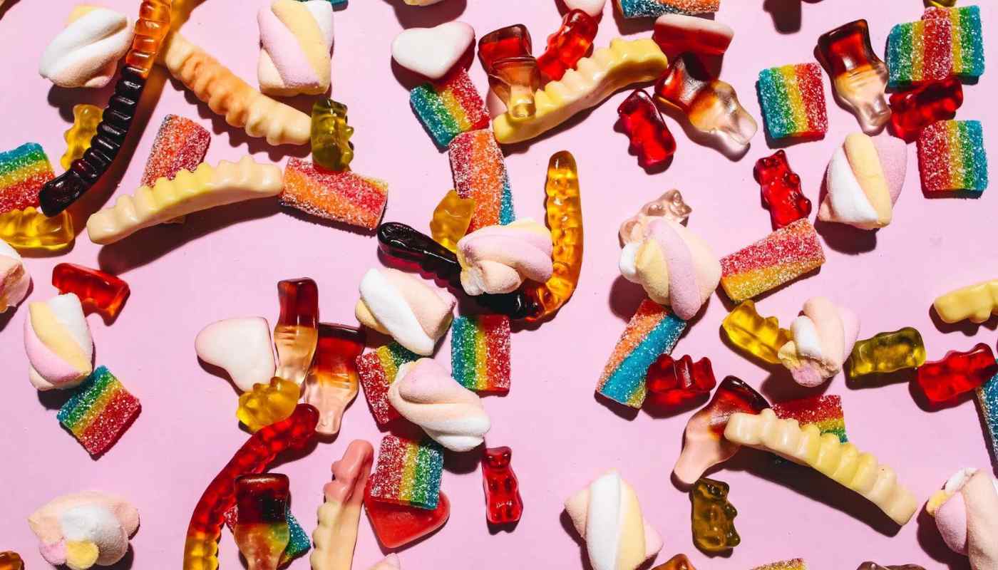Assorted gummy lollies, marshmallows and sour straps scattered on a pink background