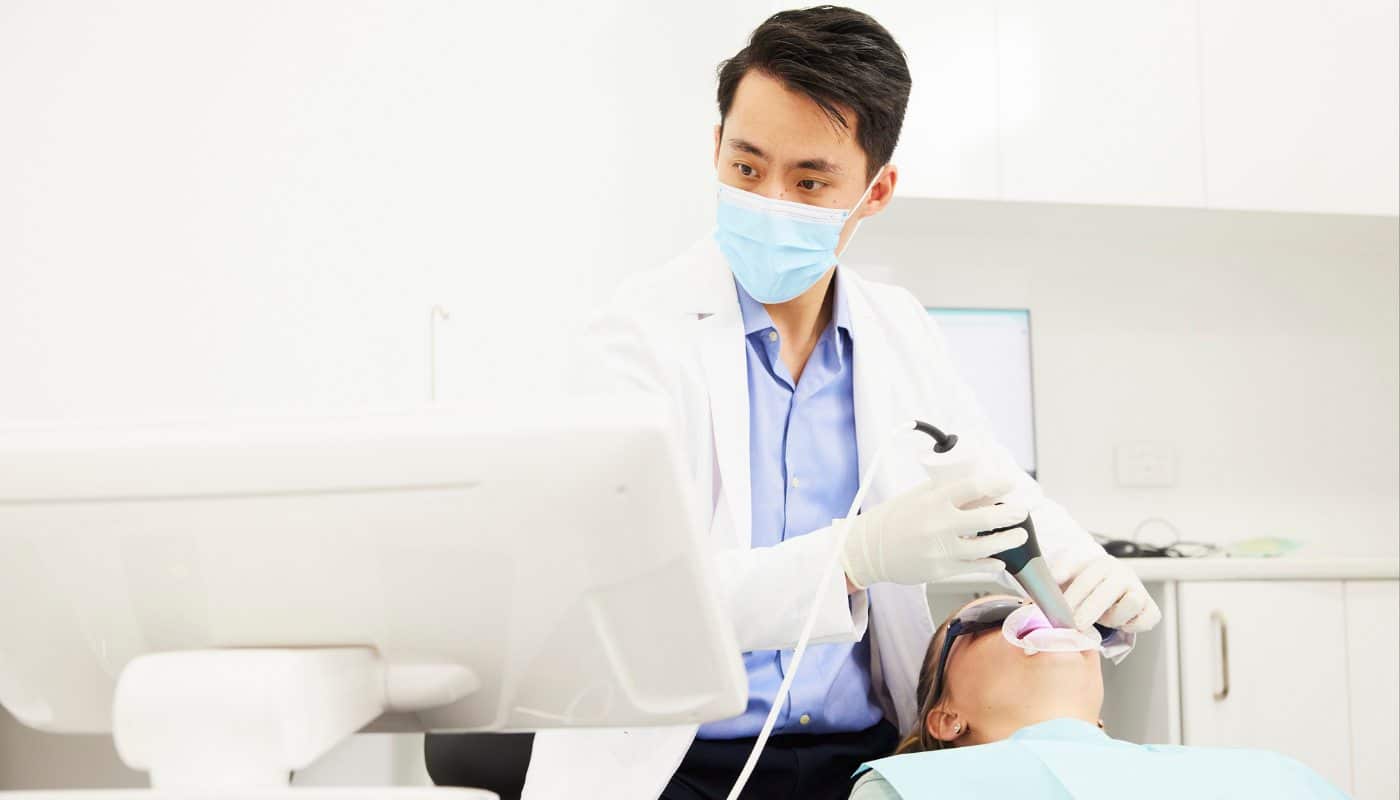 A masked dentist treating a patient reclined in the chair wearing protective glasses