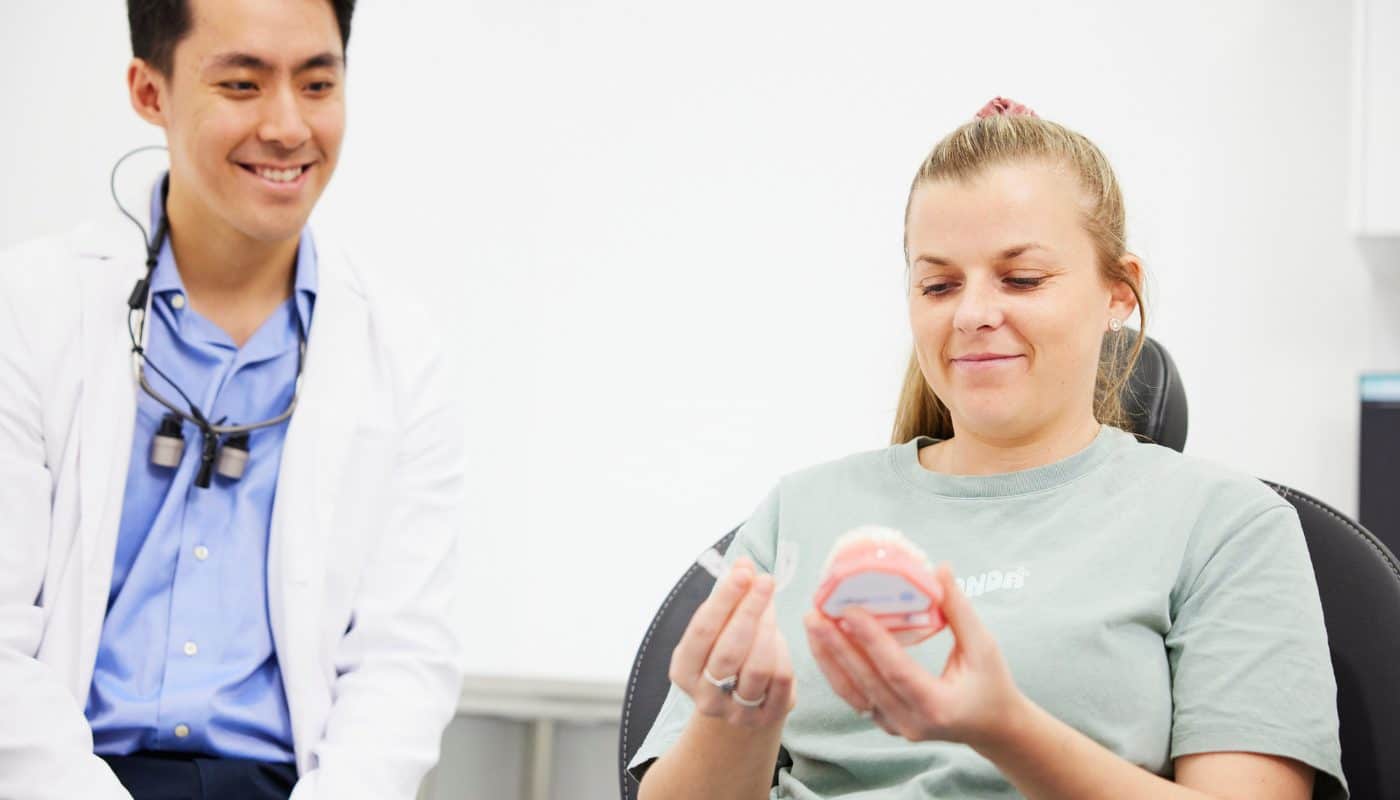 A dentist and a patient seated together, the patient holding a clear aligner and a dental model