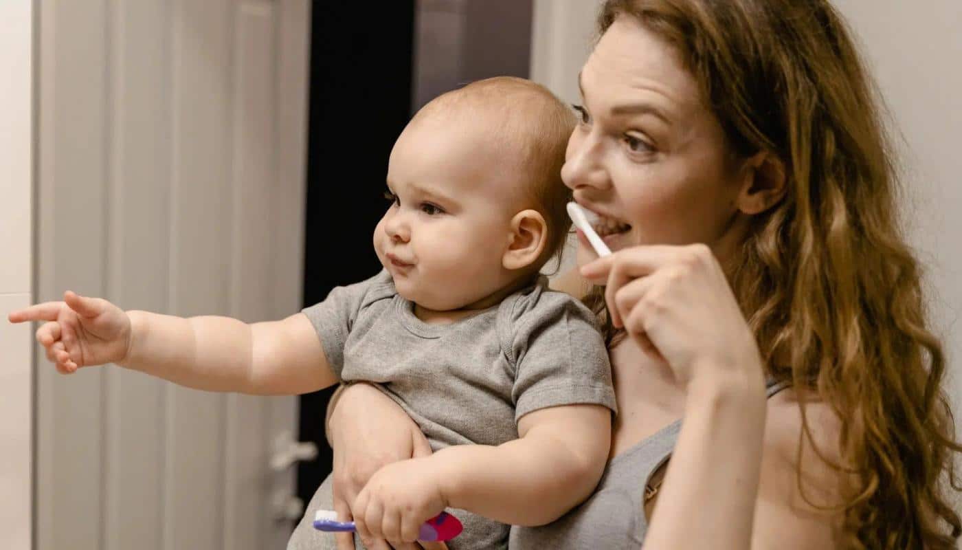 A parent brushing their own teeth while holding a baby, setting the example early