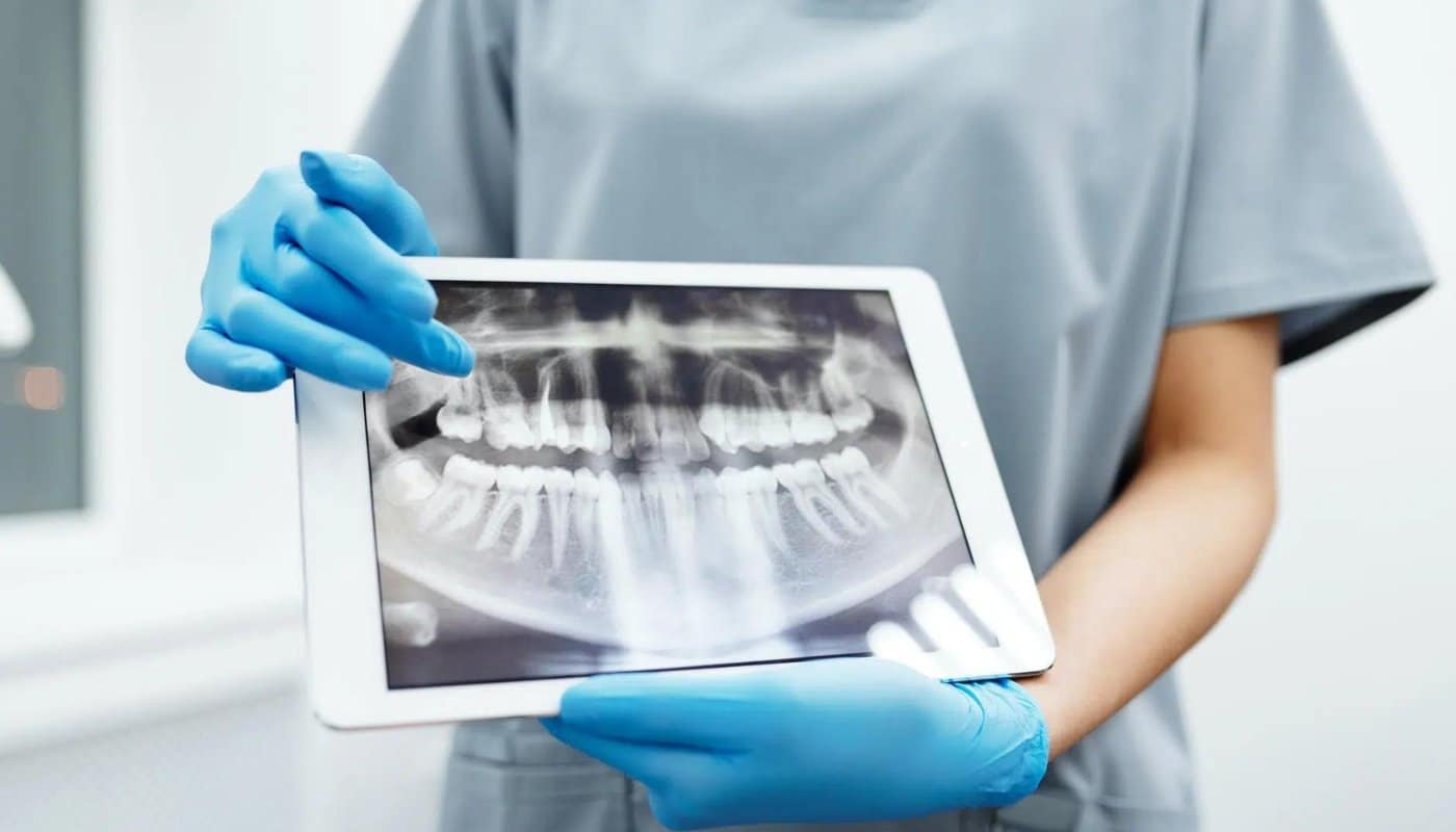A clinician in scrubs holding a tablet displaying a panoramic dental x-ray