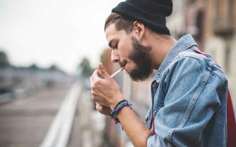 A young man in a denim jacket lighting a cigarette outdoors