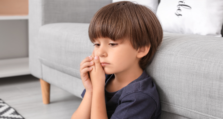 A young boy sitting on the floor beside a sofa holding his hand to his mouth
