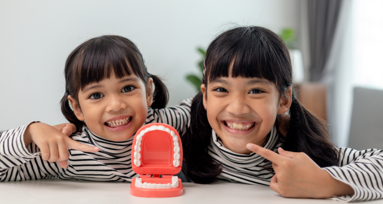 Two young girls smiling, one pointing at a model of the upper teeth on the table in front of them