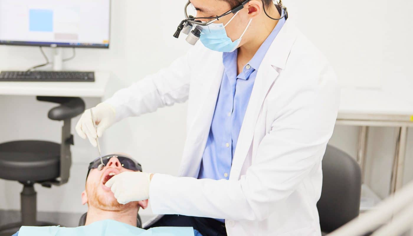 A dentist wearing magnifying loupes examining a patient reclined in the dental chair