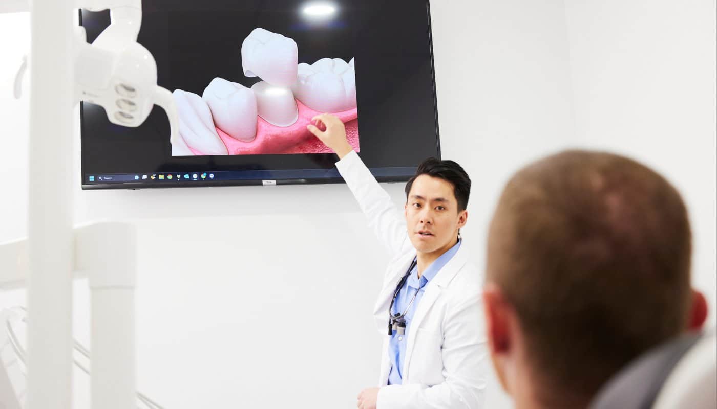 A dentist pointing to a wall-mounted screen showing a 3D animation of a dental crown while a patient watches