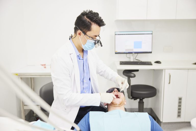 A masked dentist wearing magnifying loupes examining a patient reclined in the dental chair
