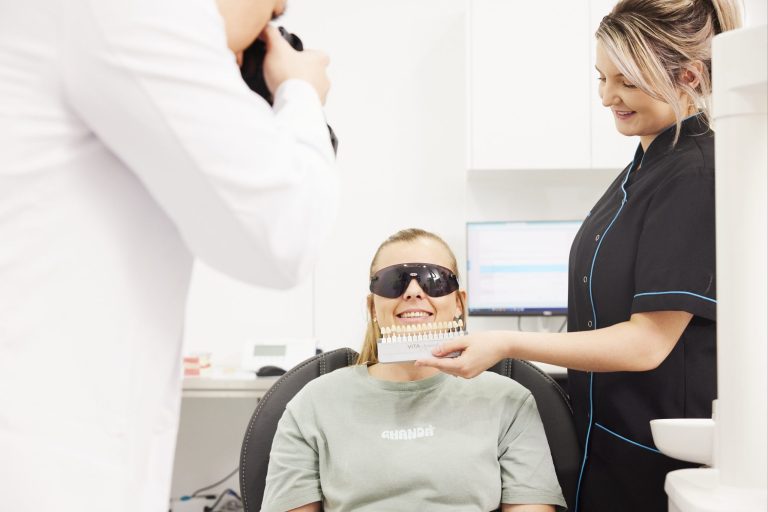 A dental assistant matching a shade guide against a patient’s teeth while the dentist photographs the smile