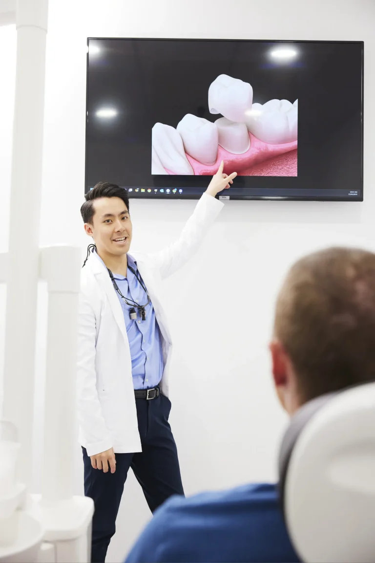 A dentist pointing to a wall screen showing a 3D animation of a dental crown while a patient watches