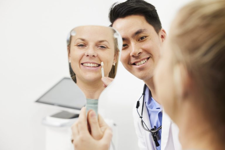 A patient holding a hand mirror and smiling while a dentist holds a tooth shade tab beside her teeth