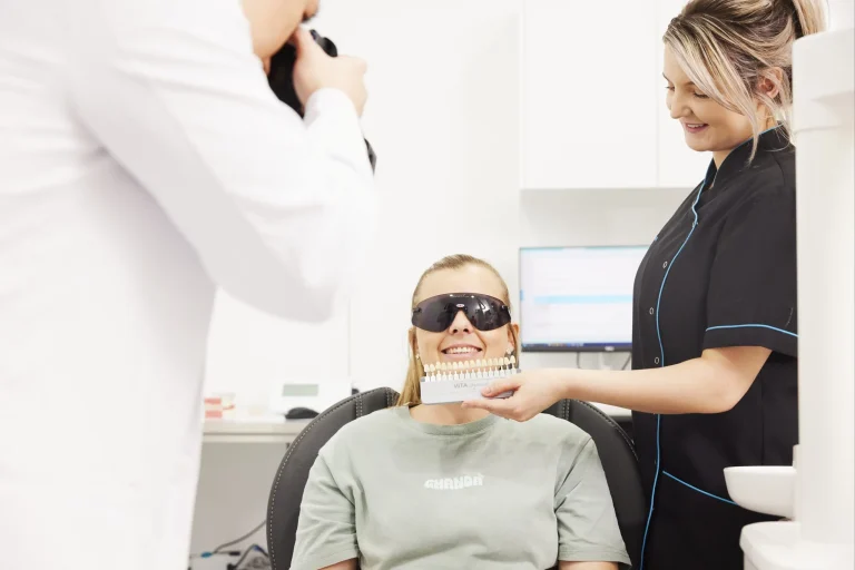 A smiling patient in the dental chair while an assistant holds a VITA shade guide beside her teeth and a clinician photographs the result