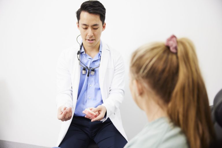 A dentist holding a clear aligner and a dental model while talking with a seated patient