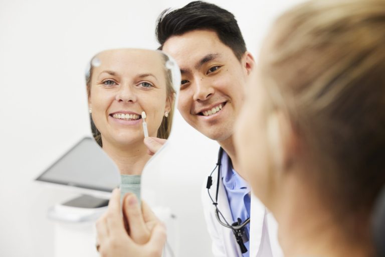 A patient smiling into a hand mirror while a dentist holds a shade tab against her front teeth