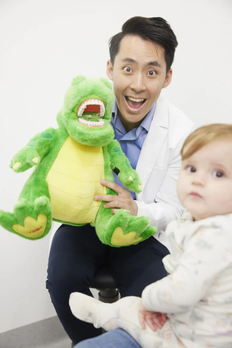 A smiling dentist holding a large crocodile puppet with teeth beside a toddler on a parent’s lap