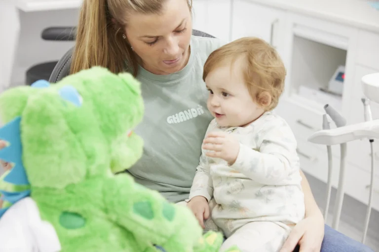 A smiling dentist holding a large crocodile puppet with teeth beside a toddler on a parent’s lap